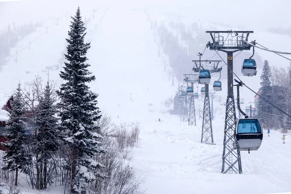 Ski lift funicular on cable car with closed cabin with blue mirrored glass on background of snow-capped mountains, blizzards, evergreen trees. Concept place for inscription, copy space