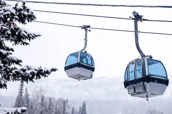 Ski lift funicular on the cable car with two closed cabins with blue mirrored glass on the background of snow-capped mountains, blizzards, evergreen trees. Concept place for inscription, copy space