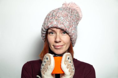 Yana doğru gülümseyen ve Studio beyaz arka plan üzerinde sıcak çay ile turuncu fincan tutan ponpon ile mor kazak pembe örgü şapka giymiş yeşil gözlü güzel genç Kızıl saçlı kız izole