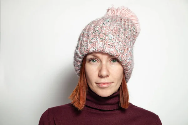 Isolated portrait of beautiful young redhead girl with green eyes in purple sweater and pink knitted hat with pompon dressed sideways smiling on white background in studio