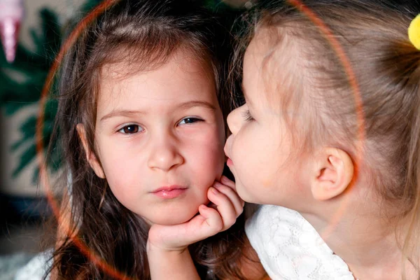 Two cute sisters of the same age lie next to the New Year tree. One kisses the other on the cheek. Concept of love and friendship of sisters and brothers, strong family and family relationships