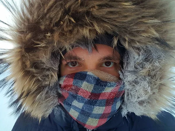 Closeup portrait young brunette man with frost covered eyebrows in warm jacket with raccoon fur hood and checkered scarf covering face. Concept of harsh weather conditions in Canada, at the North Pole