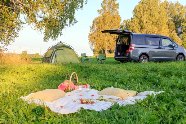 Picnic in the forest, tent and camp chairs, gray minibus with open door, blanket, wicker basket, wine glasses, snacks, fruit, bread, pillows. Concept of romantic camp date nature in sunny day