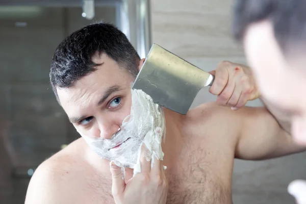 Shaving man by big knife. Concept brutal caucasian brunette men with blue eyes in white shaving foam shaves with a large kitchen cleaver, dangerous razor, barbershop, hairdresser, mirror reflection