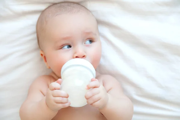 Vacker baby äta mjölk från flaska i vitt sovrum på vit bakgrund — Stockfoto