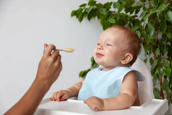 Mother feeds funny happy blond baby in blue bib from a spoon — Stock Photo, Image
