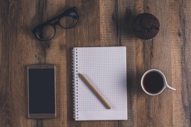 Üstten görünüm, kahve, çikolata maffin, defter, gözlük ve telefon. Çalışma alanı yüzeyi 