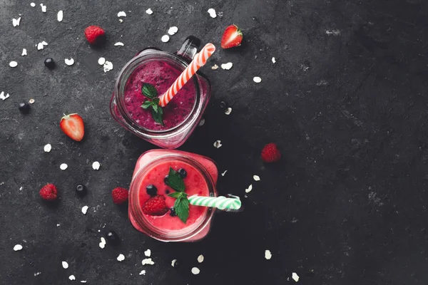 TOp view of fresh, cold smoothies made from raspberries and blueberries in glass jars with straws on dark background. Healthy diet.