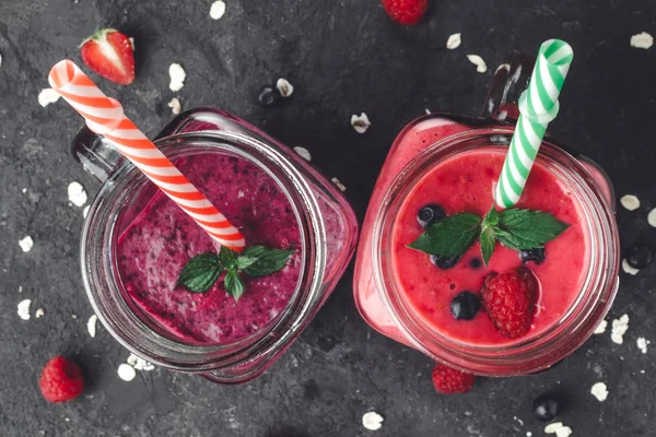 Top view of fresh, cold smoothies made from raspberries and blueberries in glass jars with straws on dark background. Healthy diet.