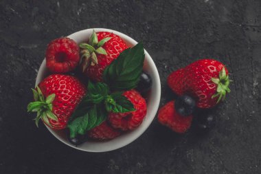 Koyu arka plan üzerine karışık çilek, ahududu ve yaban mersini küçük beyaz kase üstten görünüm.