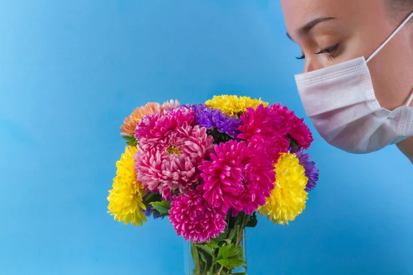 Allergy concept. Allergy to flowers bloom and pollen. A young woman in a medical mask and flowers on a blue background. Allergens, allergic reaction