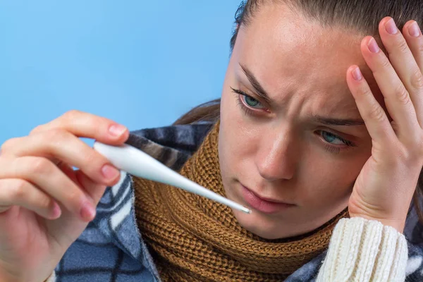 A young woman with a knitted scarf and in a warm blanket holds a thermometer in her hands and feels ill, a headache from the heat and colds Treatment of colds and flu. Weakened immunity.