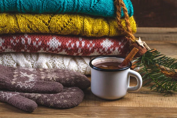 Winter, knitted, folded sweaters, warm mittens and a mug of hot cocoa with cinnamon on a wooden background. Winter and autumn clothes. Ugly Christmas Sweater