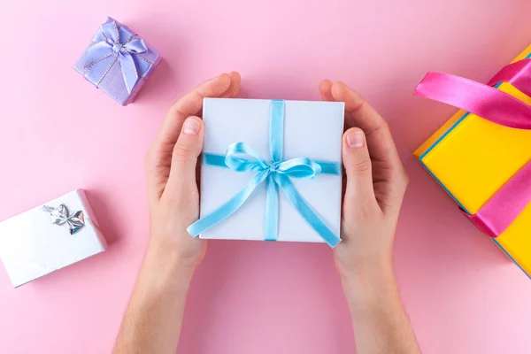 Female hands holding a small white gift box wrapped with blue ribbon. Gifts on a pink background. To give and receive gifts from loved ones.