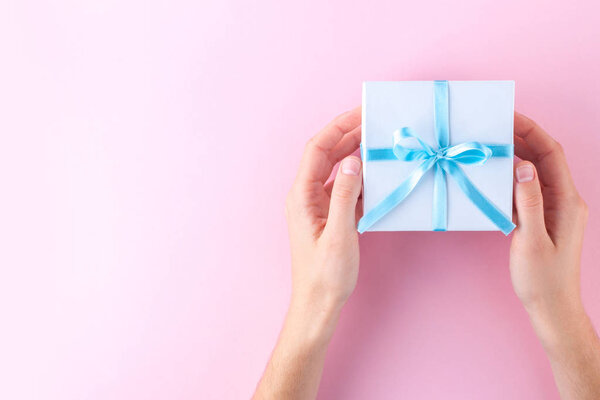 Руки женщины держат маленькую белую подарочную коробочку, завернутую голубой лентой на розовом фоне. Подарить и получить подарки от близких
. 
