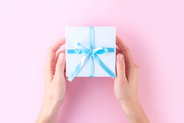 Female hands holding a small white gift box wrapped with blue ribbon on a pink background. To give and receive gifts from loved ones.