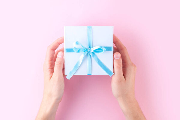 Руки женщины держат маленькую белую подарочную коробочку, завернутую голубой лентой на розовом фоне. Подарить и получить подарки от близких
. 