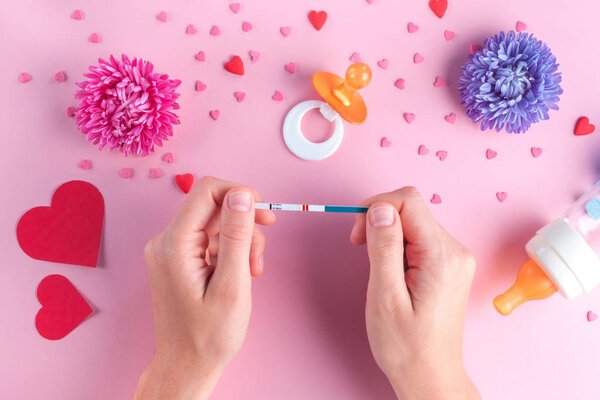 A young woman is holding a positive pregnancy test. Pregnancy concept. Happiness. Planning to become future parents. Future motherhood. Nipples, a bottle for feeding, hearts on a pink background.