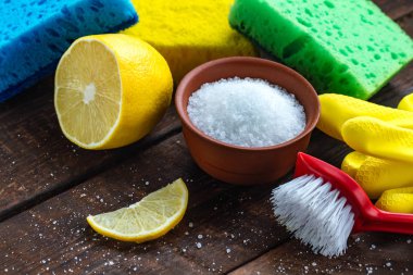 Lemon acid in a small plate, slice of lemon, a juicy lemon, sponge for washing dishes, brushes and yellow rubber gloves for cleaning the house on wooden background. Natural cleaning products