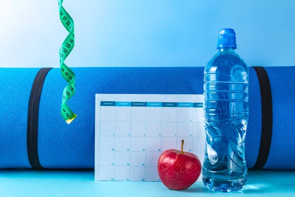 Drawing up and planning a program of sports training and diet. Sports and healthy lifestyle. An apple, a bottle of water, a measuring tape and a fitness mat on a blue background. Motivation