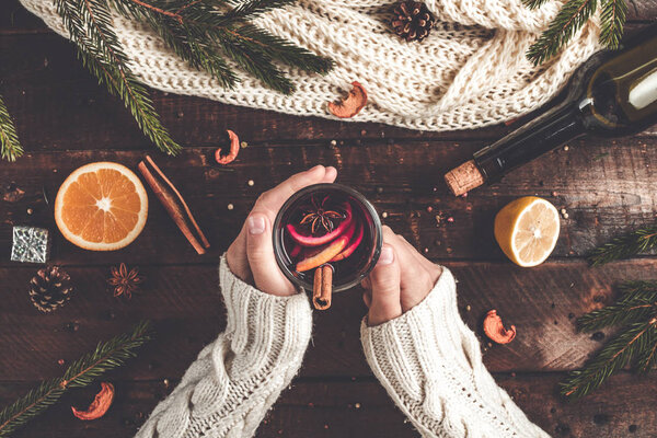 A young woman in a warm, knitted, white pullover is holding a Christmas, hot mulled wine in a glass with spices and citrus fruits. A cozy, winter evening. Winter drinks. Flat lay, top view 