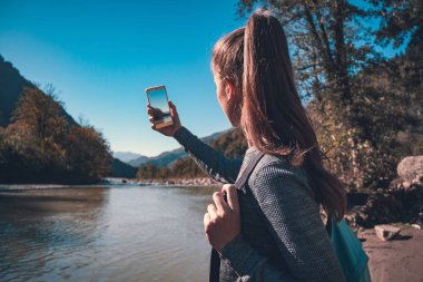 Genç kız turist mavi bir sırt çantası ile dağlarda nehir fotoğraflarını çeker. Seyahat, güzel manzara ve doğa keyfini çıkarın