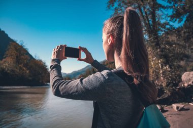 Genç kız turist mavi bir sırt çantası ile dağlarda nehir fotoğraflarını çeker. Seyahat, güzel manzara ve doğa keyfini çıkarın