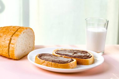 Somun ile yayılan çikolatalı hamur beyaz tabakta ve bir tablo üzerinde bir taze pişmiş ekmek parçaları. Tatlı, fındık yapıştırın taze ekmek ve bir bardak süt ile kahvaltıda.