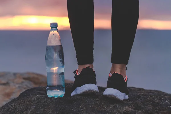 夕暮れ海の背景の上に水のボトルの横にある岩の上に立ってスニーカーの女性 スポーツやアクティブなライフ スタイル — ストック写真