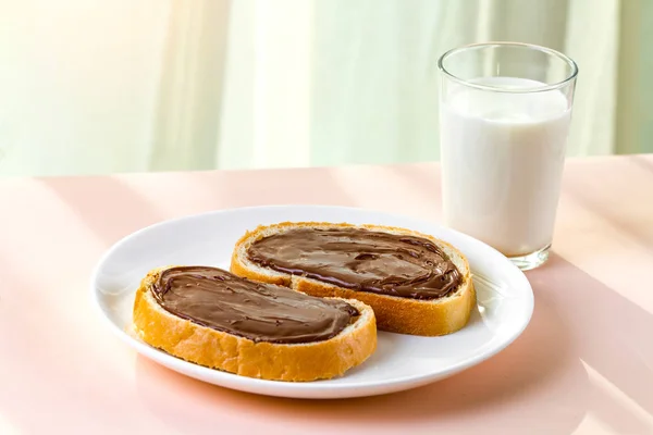 A pieces of loaf with spread chocolate paste on a white plate. Sweet, chocolate sandwiches with nut paste and a glass of milk for breakfast.