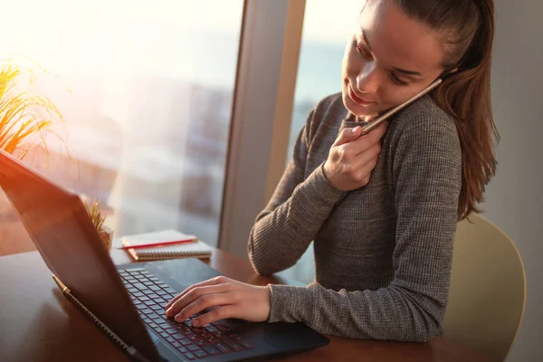 オンライン作業と携帯電話で話しているラップトップを使用して 職場で座っている美しい若いビジネス女性 — ストック写真