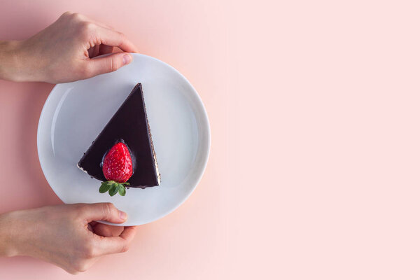 A slice of sweet strawberry cake with dripping chocolate glaze in a white plate in hands on a pink background. Sweet dessert. Copy space 