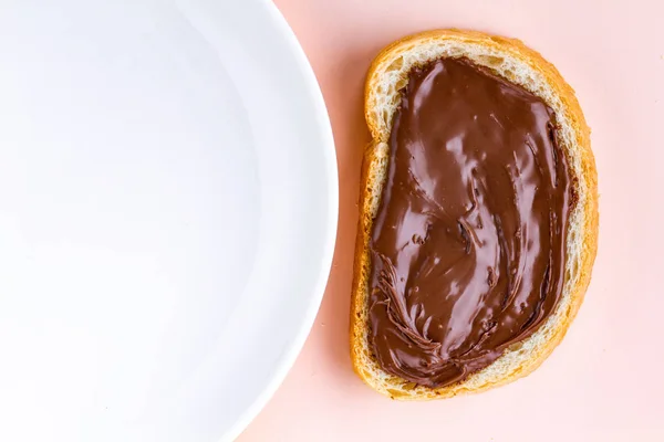 Piece of loaf with chocolate paste and a plate on a pink background. Sandwich with nut paste on a breakfast. Top view. Copy space