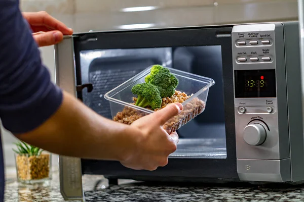 Using the microwave oven to heat food. Woman\'s hand puts plastic container with broccoli and buckwheat in the microwave