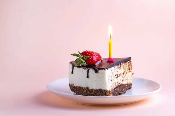 Strawberry cake with cream, dripping chocolate glaze and a lit candle in a plate on a pink background for birthday and holiday celebrations. Copy space. Make a wish and blow out the candle on the birthday cake