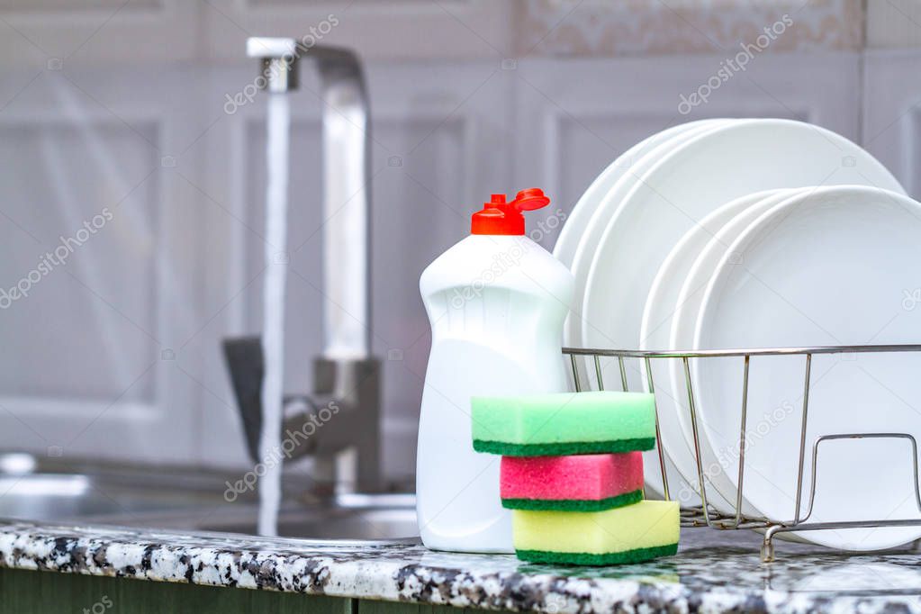 Different clean plates in dish drying rack, dish sponges and dishwashing detergent on the table on kitchen counter. Washing dirty dishes