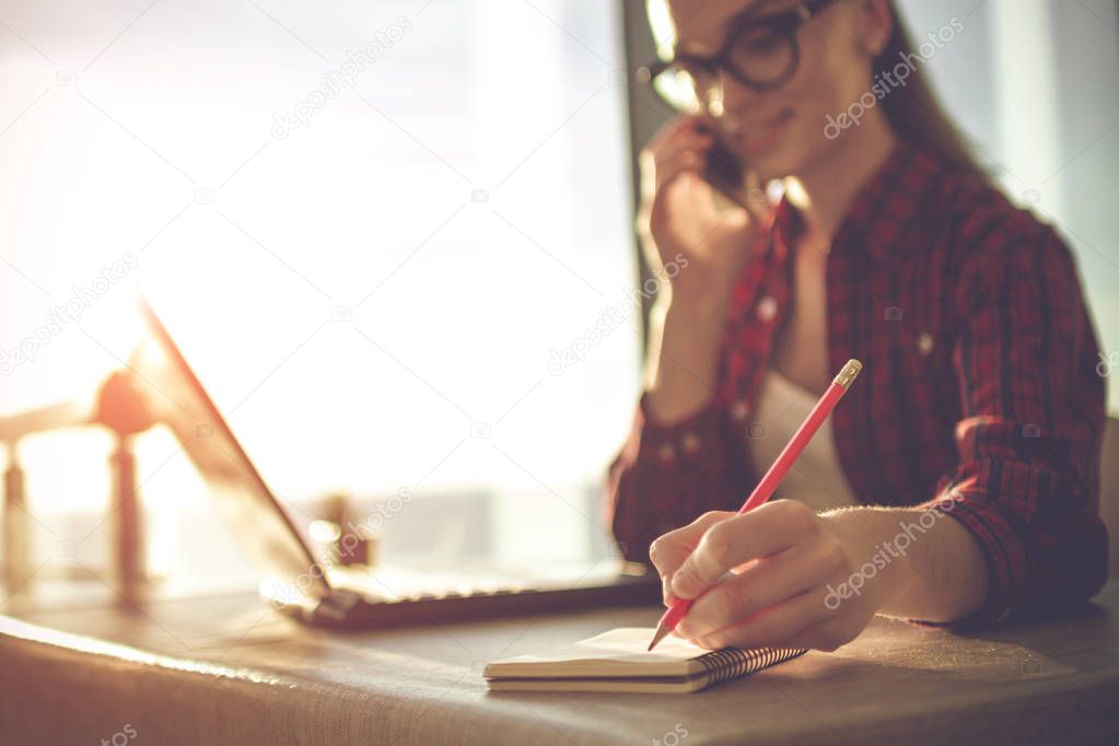 Young, attractive business woman talking on the phone and takes a note in her notebook while working online on a laptop at home.