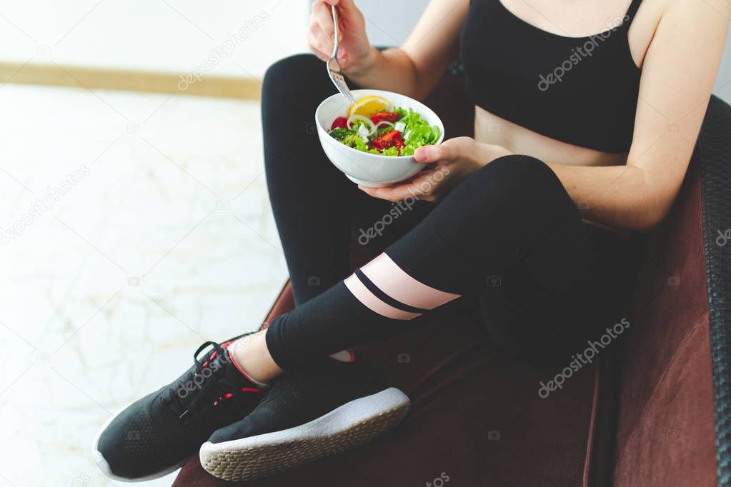 Fitness woman in sneakers and sportswear is resting and eating a healthy, fresh salad after a workout. Fitness and healthy lifestyle concept. 