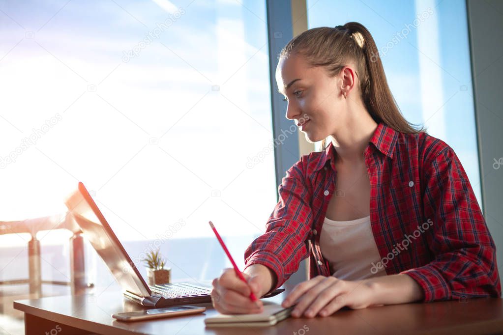 Young, attractive business woman takes a note in her notebook while working online on a laptop at home. 