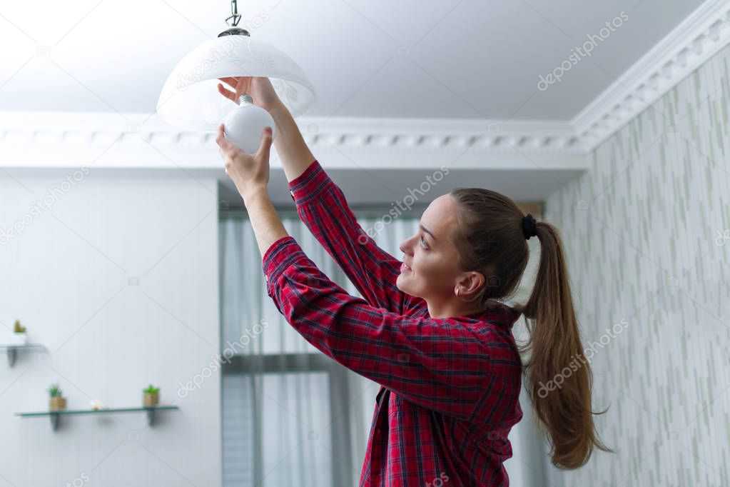 Young, attractive housewife makes the replacement of the used LED light bulb in the chandelier in the room 