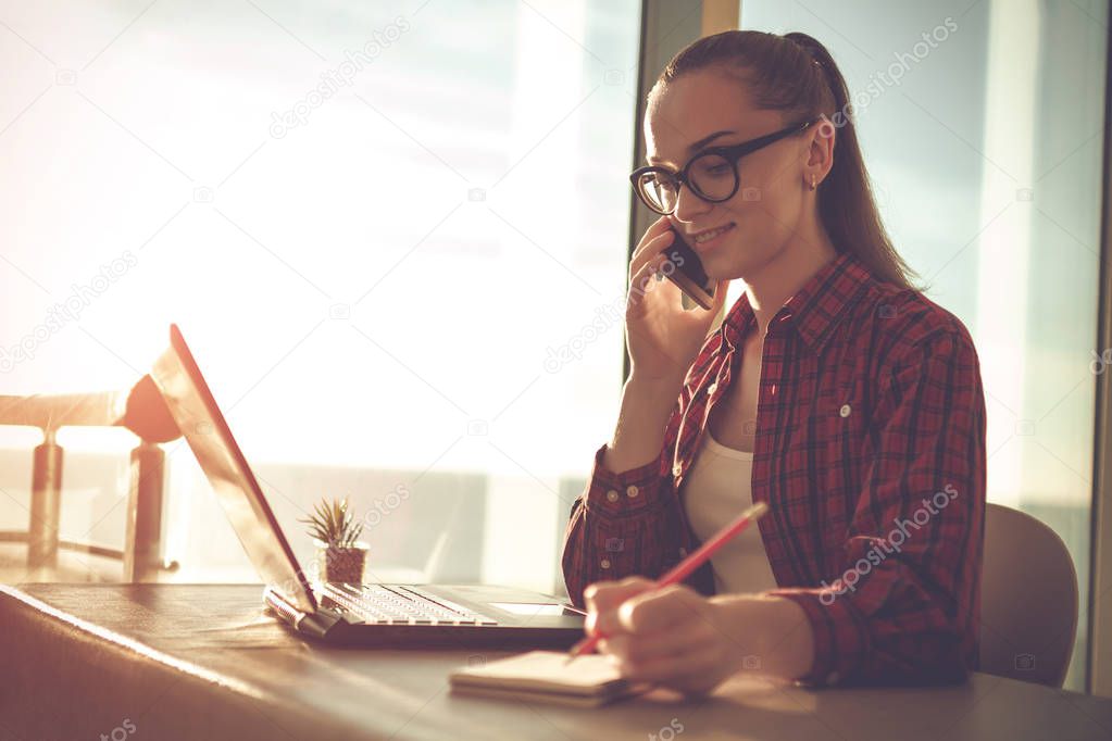 Young, attractive business woman talking on the phone and takes a note in her notebook while working online on a laptop at home.