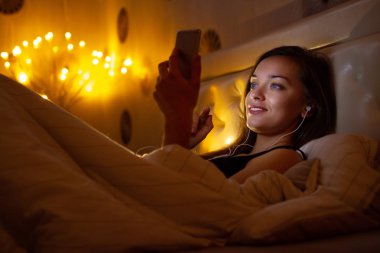 Yatmadan önce kulaklıklı genç bir kadın internette müzik dinliyor ve sohbet ediyor. Sohbet gecesi ve sörf telefonu. Sosyal ağlar ve telefon bağımlılığı. 