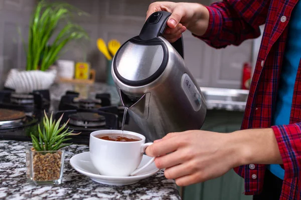 A person makes tea using boiling water from an electric kettle in kitchen at home. Time for breakfast