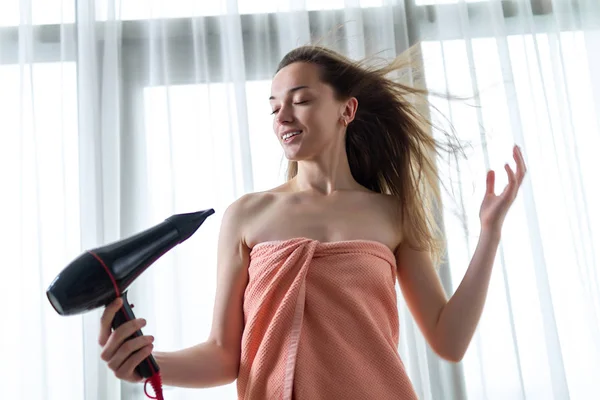 バスタオルの美しい若い茶色の髪の女性は 部屋でシャワーの後に彼女の長い髪を乾燥させるためのヘアドライヤーを使用しています ヘアケア — ストック写真