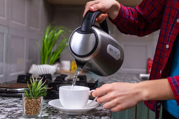 A person makes tea using boiling water from an electric kettle in kitchen at home. Time for breakfast