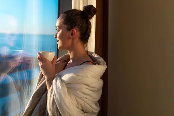 毛布に包まれた非常に若い女性が窓の近くに立っているとサンシャインで最初の朝のコーヒーを楽しんでいます 早く目覚め 新しい日の始まり — ストック写真