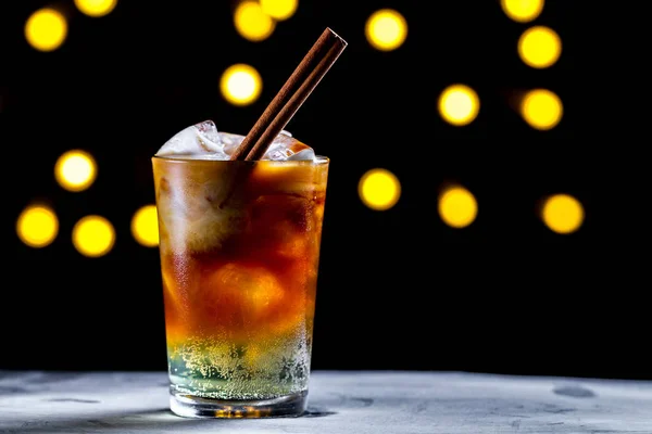 Refreshing cocktail with ice cubes, cinnamon and soda bubbles on the background of lights. Ice coffee. Cold and carbonated drinks