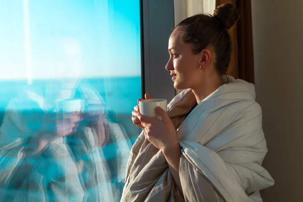 毛布に包まれた非常に若い女性が窓の近くに立っているとサンシャインで最初の朝のコーヒーを楽しんでいます 早く目覚め 新しい日の始まり — ストック写真
