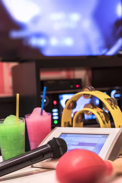 Black microphone in karaoke club, with remote controller, melon and strawberry soda drinks, yellow tambourine and screen for singing music on stage party.