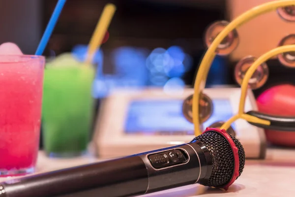 Black microphone in karaoke club, with remote controller, melon and strawberry soda drinks, yellow tambourine and screen for singing music on stage party.